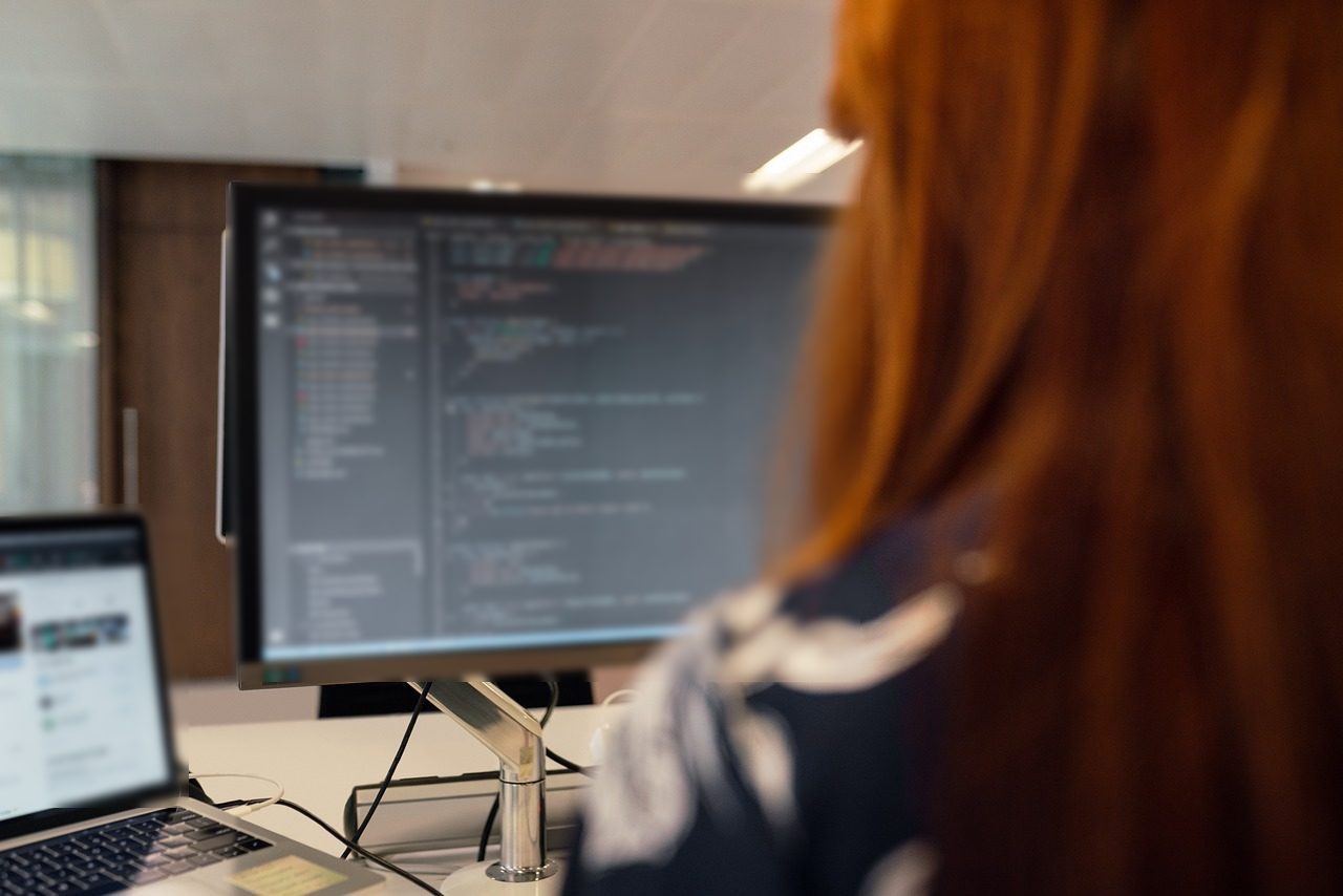 Specialistă văzută din spate, cu editorul de cod pe monitor și pagina site-ului pe laptop